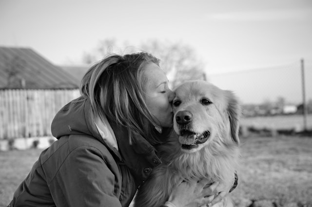 A black-and-white photo of a woman kissing a golden retriever on the cheek. The woman is wearing a jacket, and the dog looks content with its mouth slightly open. They are outdoors near a wooden shed and a wire fence.
