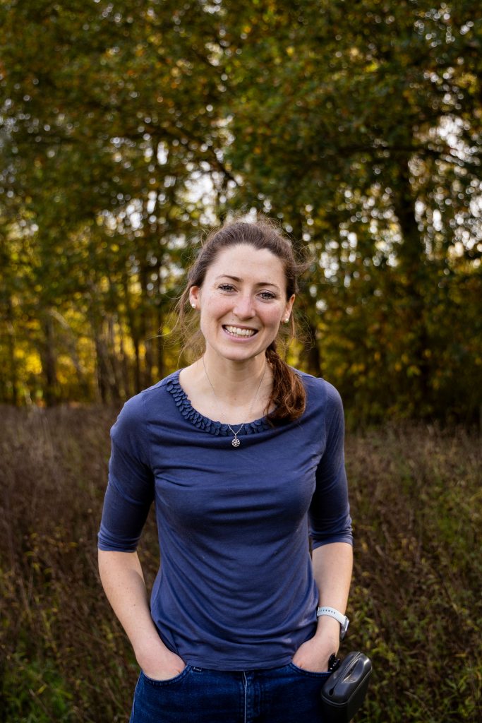 A woman smiling in a wooded area, with her hands in her pockets.